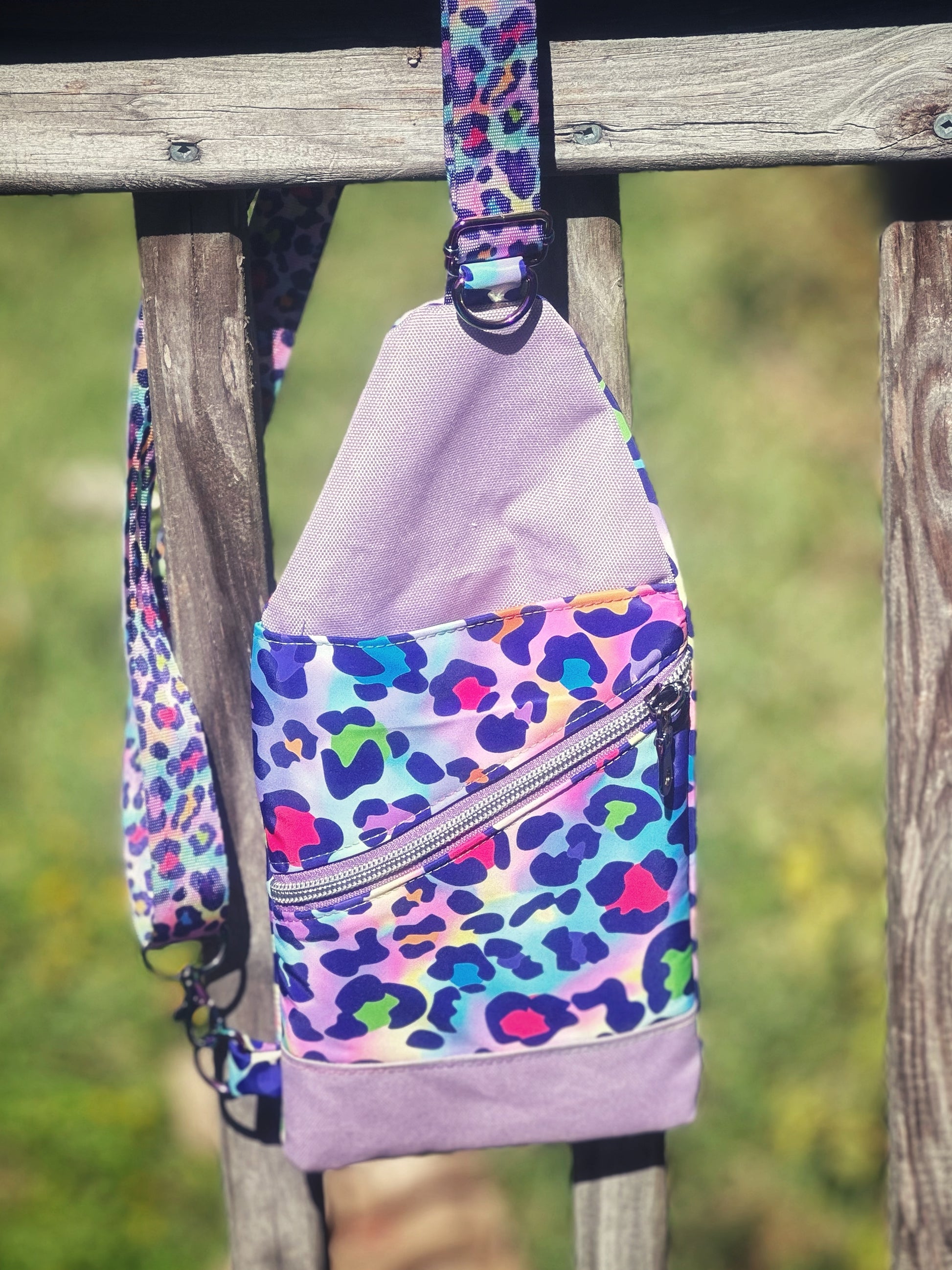 Front view of a handmade sling bag featuring a bold rainbow leopard print in neon pink, blue, green, and purple against a pastel background. The bag has a diagonal zipper pocket with a silver zipper and is accented with light purple canvas on the top and bottom. The strap is made from matching leopard webbing. The bag hangs on a rustic wooden fence with a grassy yard in the background.