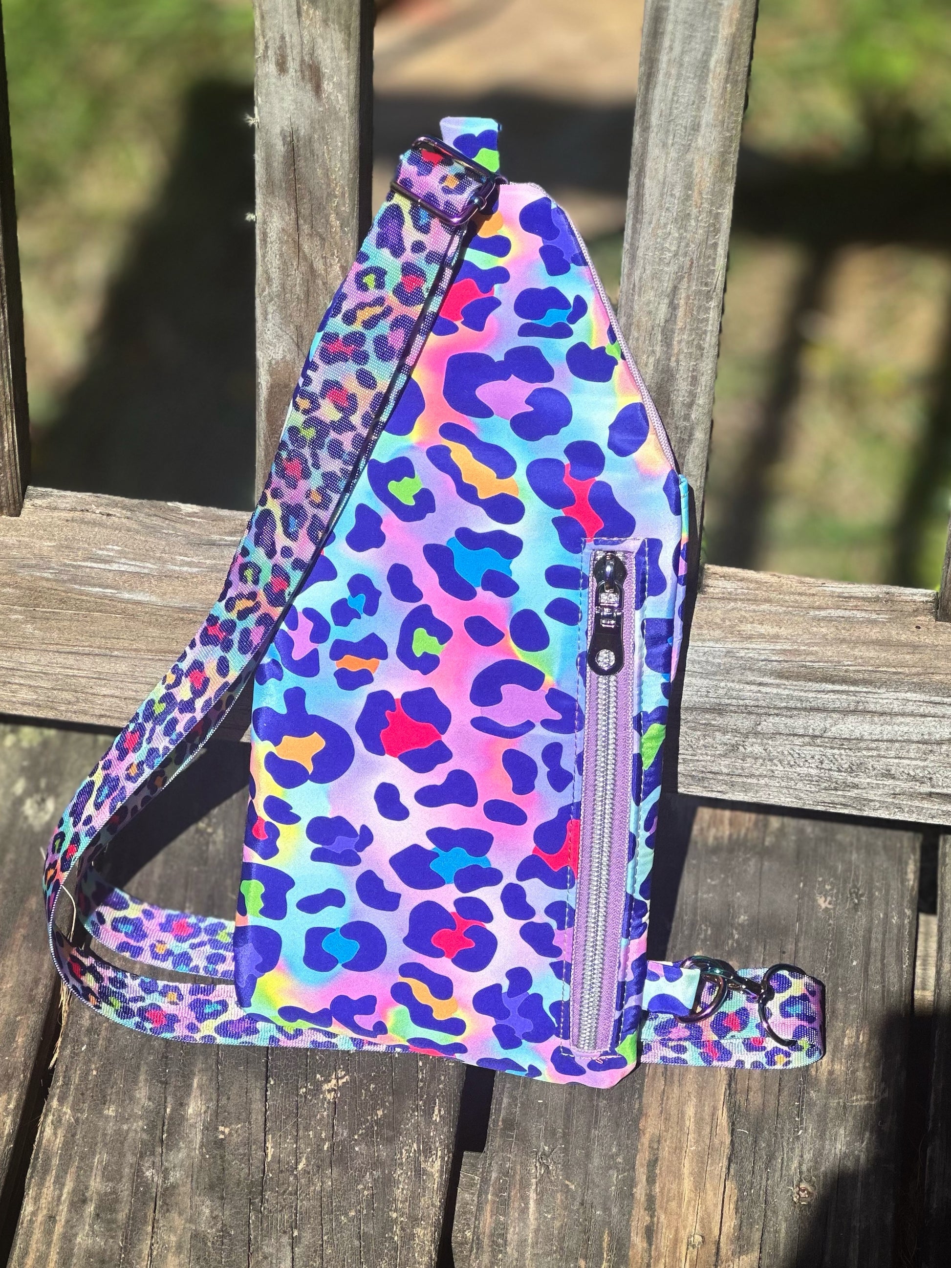 Back view of a handmade sling bag in a vibrant rainbow leopard print. The fabric features bold spots in pink, blue, purple, yellow, and green on a pastel gradient background. A vertical zipper pocket with a light lavender zipper and matching rainbow leopard strap is visible. The bag is displayed leaning on a weathered wooden deck in bright sunlight, with green grass in the blurred background.