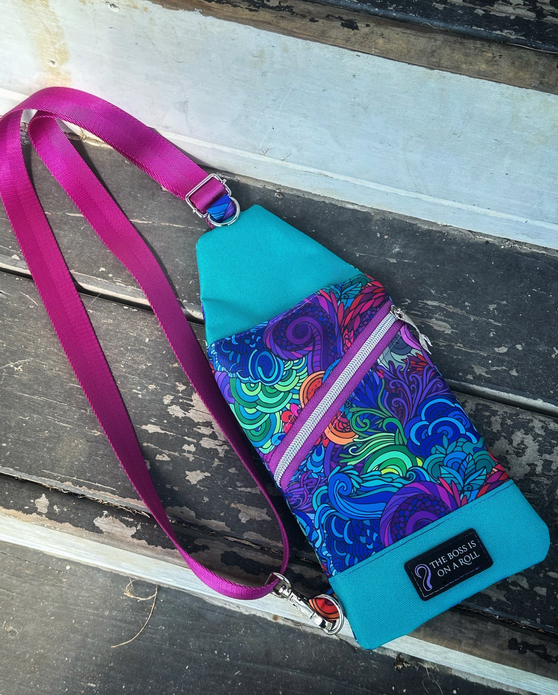 A handmade sling bag featuring a vivid under-the-sea inspired print in swirling blues, purples, and greens, reminiscent of sea grass and coral reefs. The front has a diagonal purple zipper with a silver mermaid tail charm on the pull. The top and bottom panels are blue canvas, and a black patch near the base reads “The boss is on a roll,” alongside a curled tentacle graphic. The crossbody strap is magenta with silver hardware.