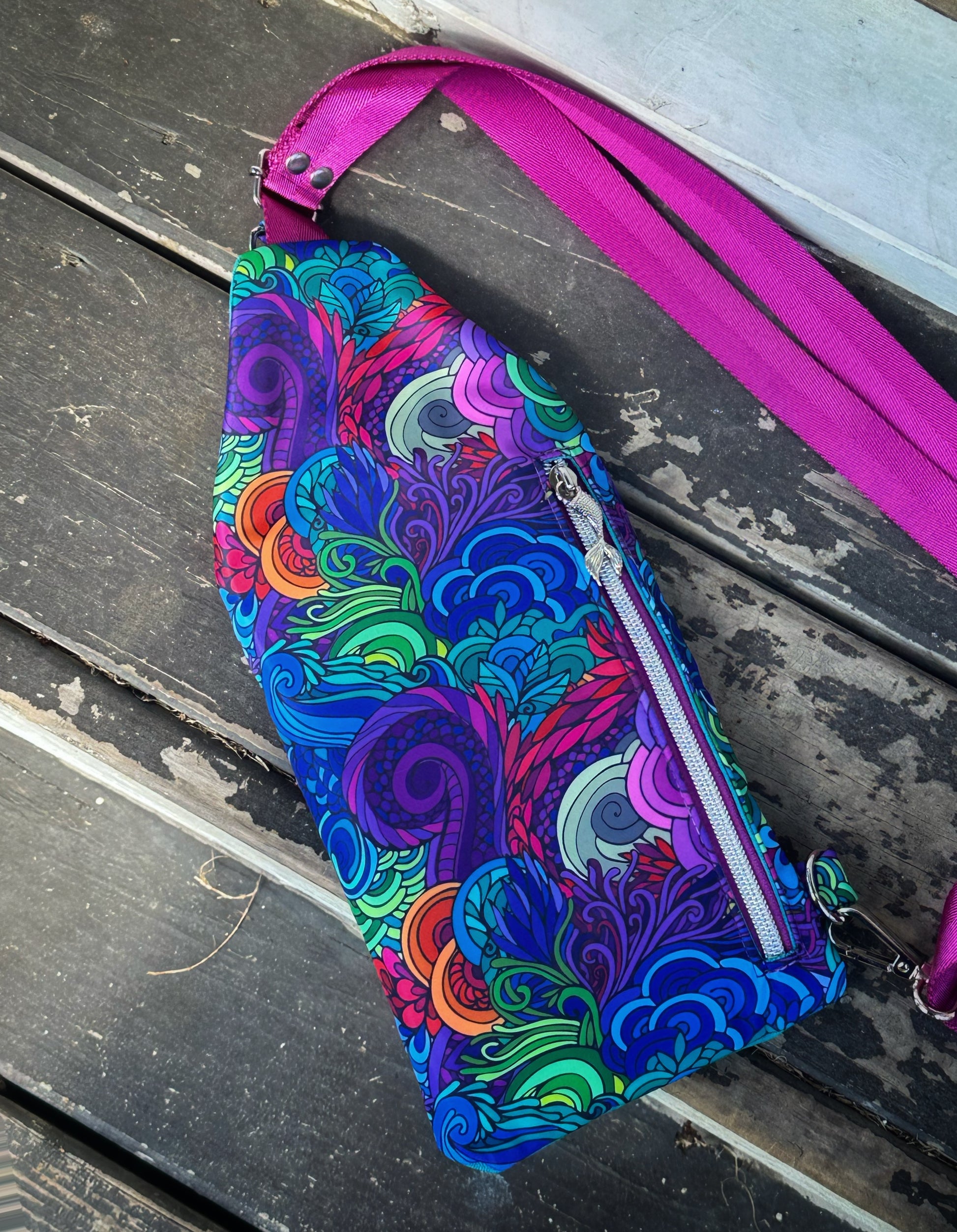 Back view of a handmade sling bag featuring a swirling undersea-inspired print in bright blues, purples, greens, and teals, resembling sea grass and coral reef textures. A vertical purple zipper runs down the right side, topped with a silver mermaid tail charm. The magenta crossbody strap is visible at the top right corner.