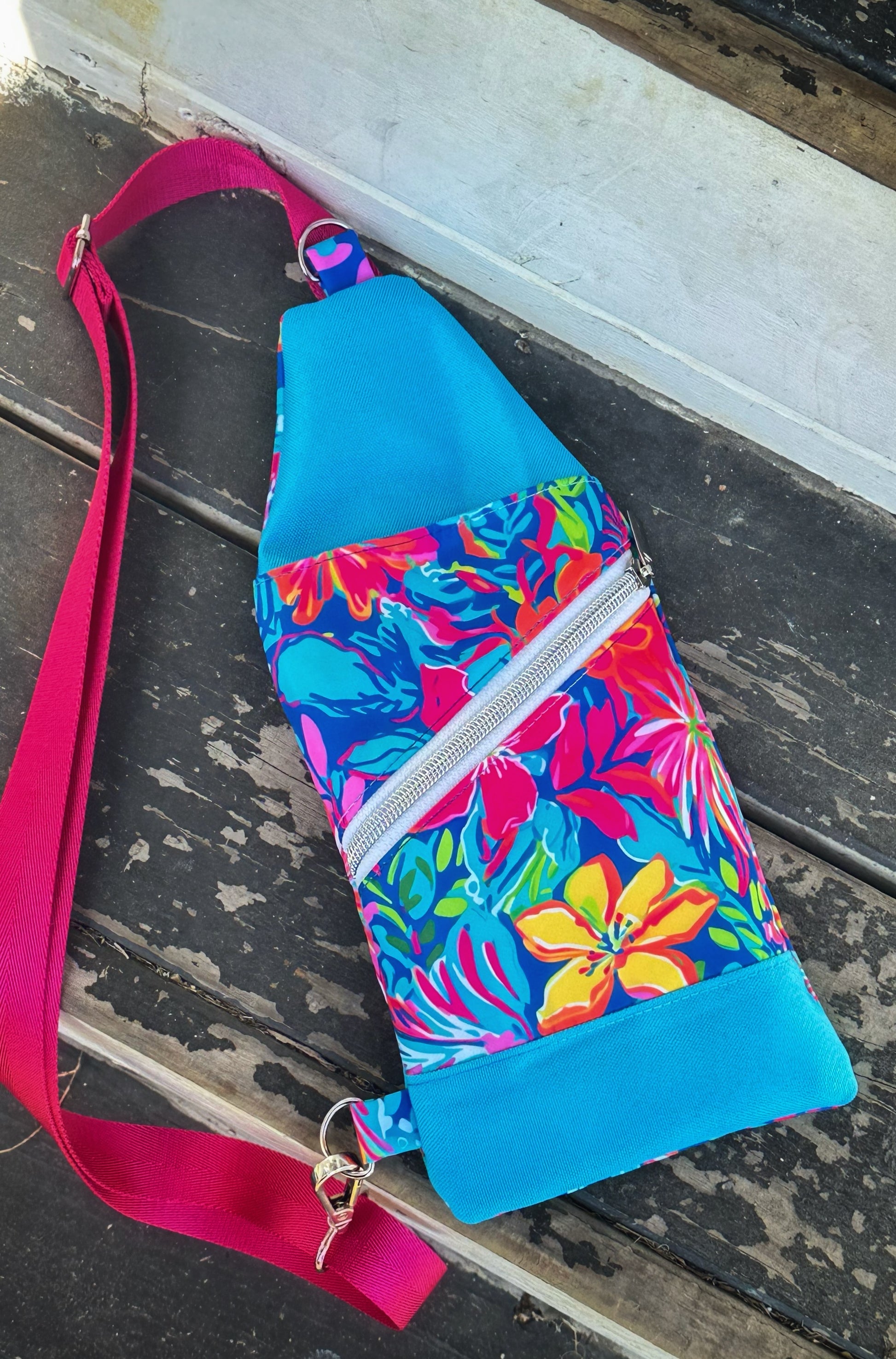 Front view of a handmade sling bag with a bright tropical floral print in bold pinks, yellows, blues, and greens. The top and bottom panels are a vivid sky blue, and a white diagonal zipper crosses the front. A hot pink crossbody strap is attached at the upper middle and lower left corner.