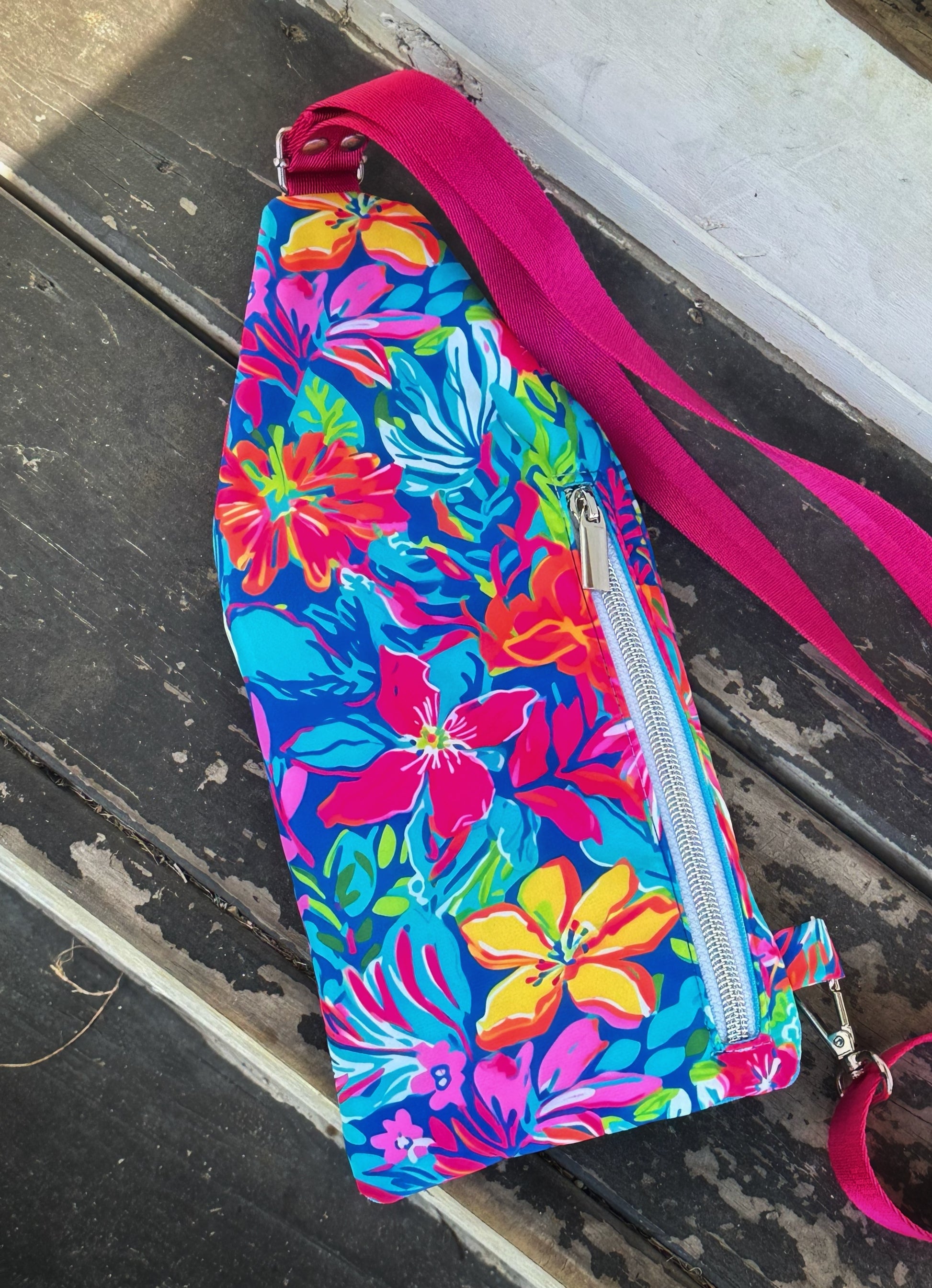Back view of a handmade sling bag featuring a vibrant tropical floral print in bold pink, red, yellow, blue, and green. A silver vertical zipper pocket runs down the right side. The hot pink strap attaches at the top middle and bottom left corner. The background is a weathered wooden surface.