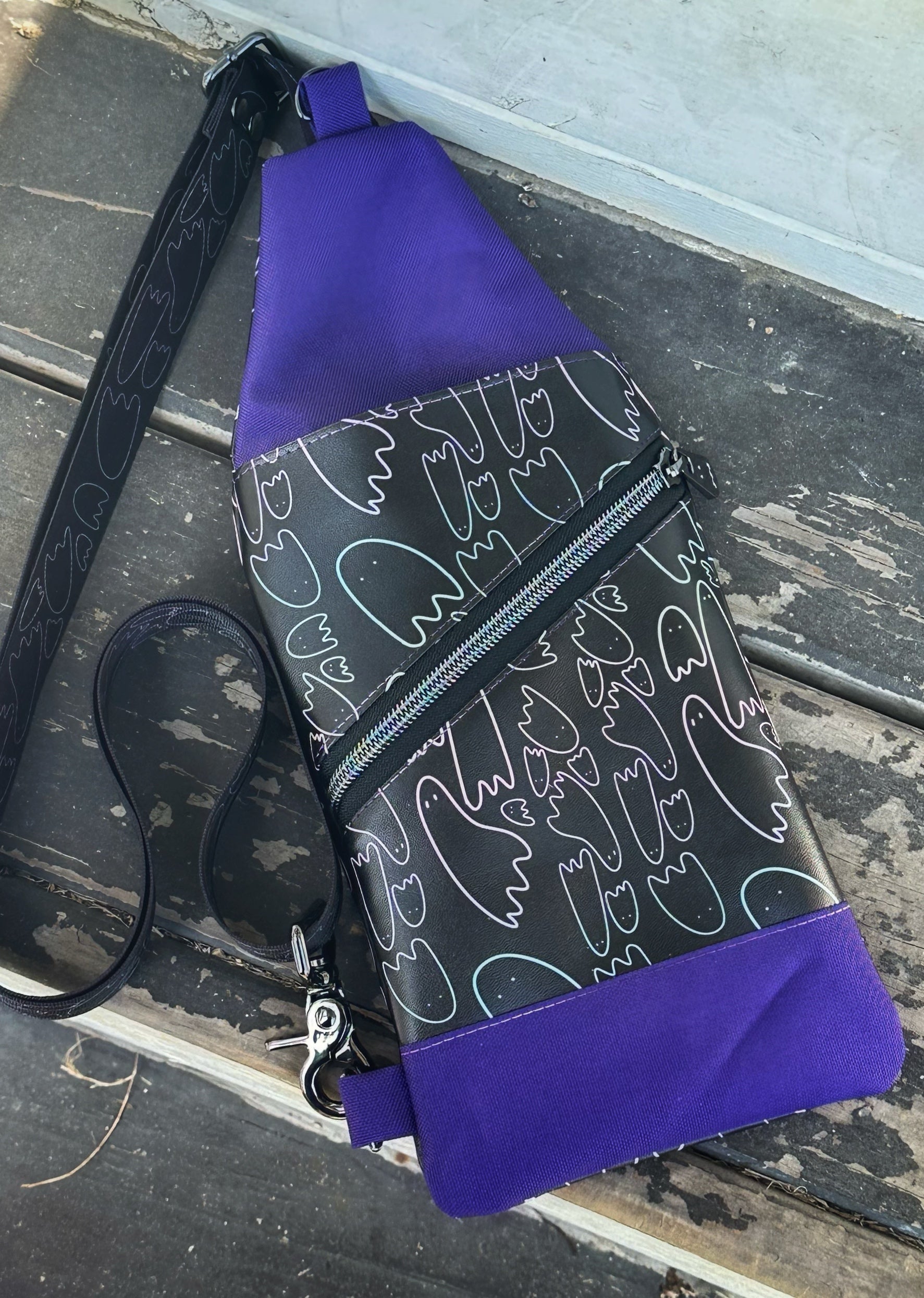 A handmade sling bag lies flat on a worn wooden surface. It features black fabric with pastel ghost outlines, bold purple top and bottom panels, a diagonal front zipper, and a black crossbody strap with ghost designs.