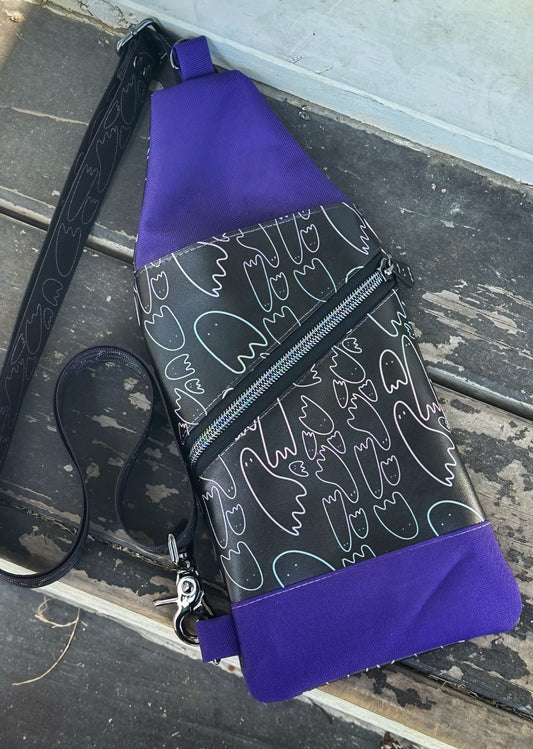 A handmade sling bag lies flat on a worn wooden surface. It features black fabric with pastel ghost outlines, bold purple top and bottom panels, a diagonal front zipper, and a black crossbody strap with ghost designs.