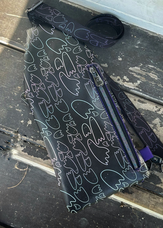 A handmade sling bag lies flat on weathered wooden planks. The bag is made from black material patterned with pastel ghost outlines in pink, purple, blue, and mint. A vertical zipper runs down the center right side of the bag, featuring a ghost-detailed zipper pull. The strap, also patterned with ghost outlines, is partially coiled near the top.