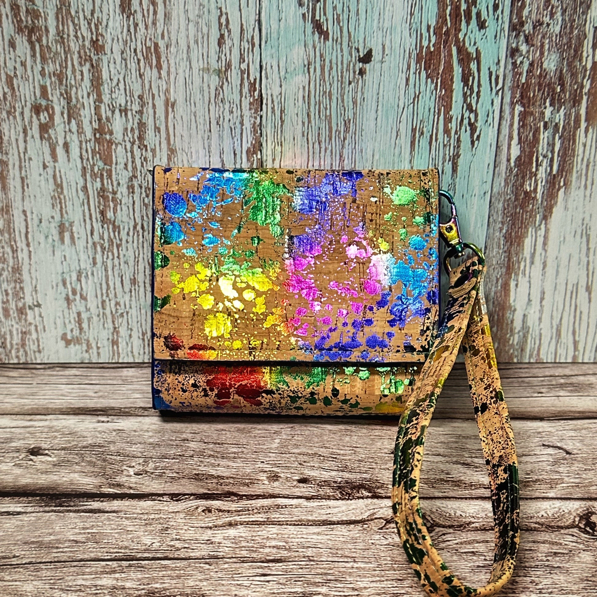 A compact handmade wallet crafted from natural cork material with a bold, colorful splatter paint design in rainbow hues—yellow, green, blue, purple, pink, and red. The front flap folds over neatly and is secured with a hidden snap. Attached is a matching cork wrist strap with coordinating paint splatter. The wallet is displayed upright against a distressed aqua and woodgrain backdrop on a rustic wooden surface.