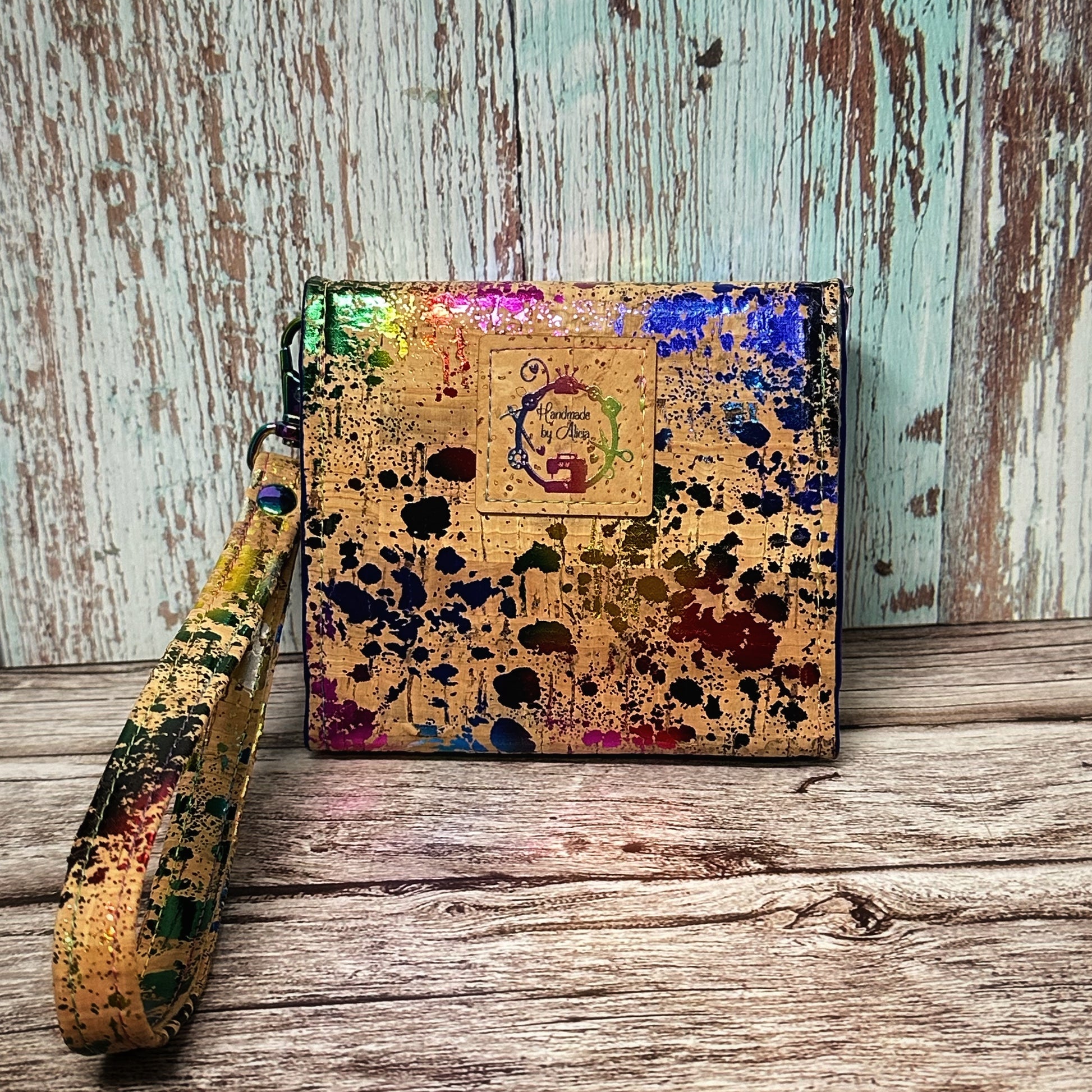 The back view of a handmade cork wallet featuring a vivid rainbow splatter paint design on natural cork. A square cork tag is centered near the top and stamped with the logo “Handmade by Alicia” surrounded by sewing bits and bobs and a small sewing machine. The detachable wrist strap matches the wallet’s paint-splatter cork pattern. The background includes a weathered teal and wood plank backdrop and rustic wooden surface.