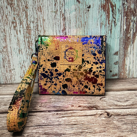 The back view of a handmade cork wallet featuring a vivid rainbow splatter paint design on natural cork. A square cork tag is centered near the top and stamped with the logo “Handmade by Alicia” surrounded by sewing bits and bobs and a small sewing machine. The detachable wrist strap matches the wallet’s paint-splatter cork pattern. The background includes a weathered teal and wood plank backdrop and rustic wooden surface.