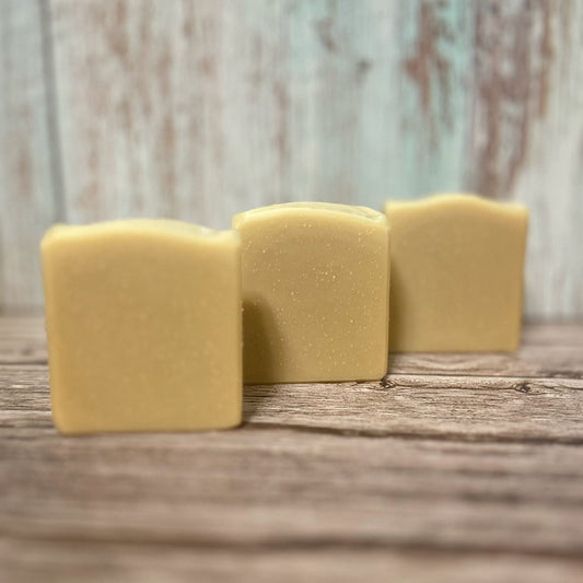 Three handmade soap bars in a soft, golden yellow tone are lined up diagonally on a rustic wooden surface. Each bar has a slightly curved top and a smooth, natural finish. The background features distressed pale teal and white wood paneling.