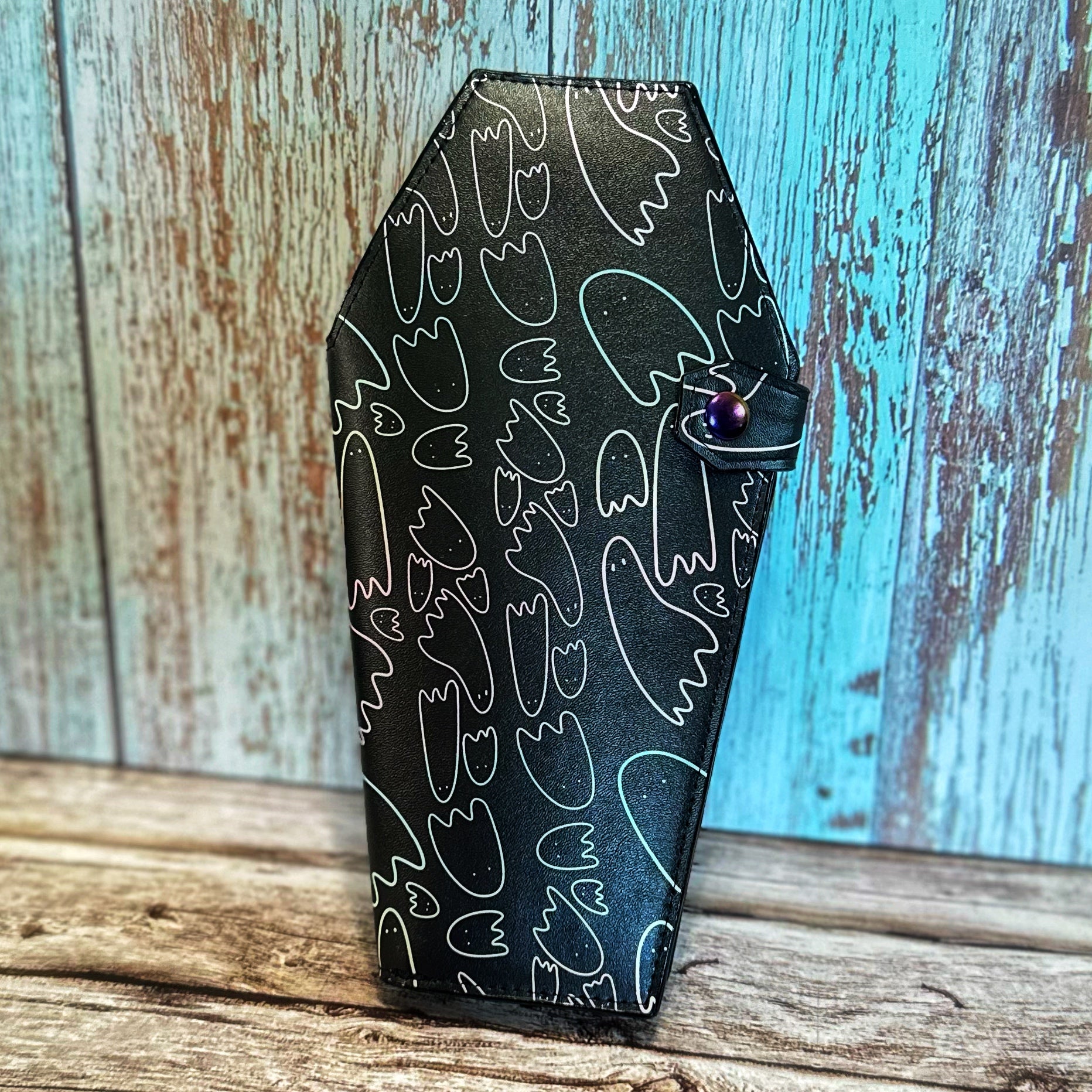 Front view of a tall, handmade coffin-shaped wallet crafted from black vinyl with whimsical pastel-outlined ghost doodles. The wallet is secured with a coffin-shaped tab and a rainbow snap closure. The background is rustic wood paneling in shades of teal and cream.
