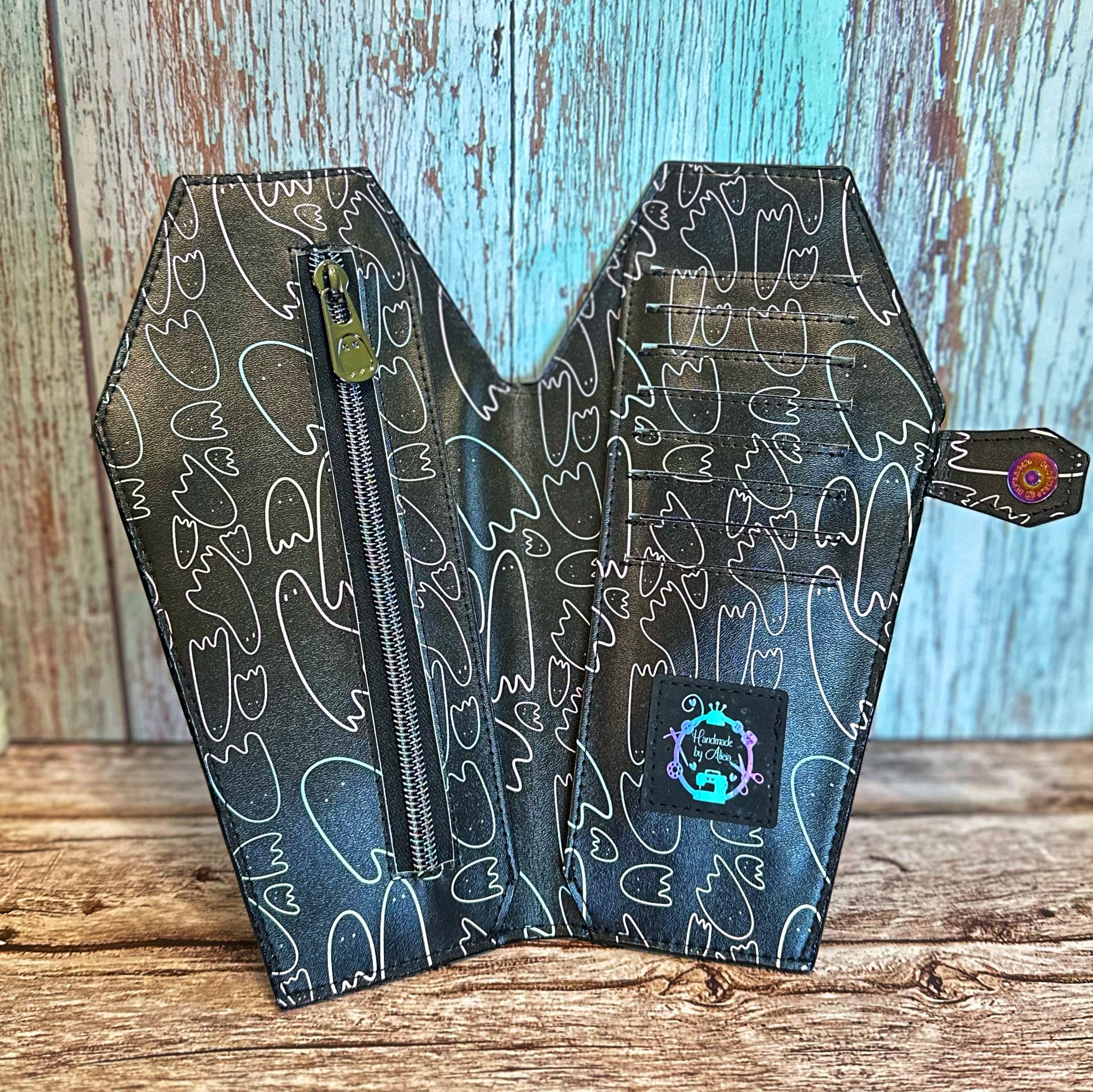 Interior view of a tall, handmade coffin-shaped wallet made from black vinyl with pastel-outlined ghosts. The wallet features eight card slots, a zippered coin pocket with a gunmetal ghost-shaped zipper pull, and two full-length cash slots. A cork 'Handmade by Alicia' logo tag is sewn below the card slots, and the closure tab with rainbow snap is visible on the right.