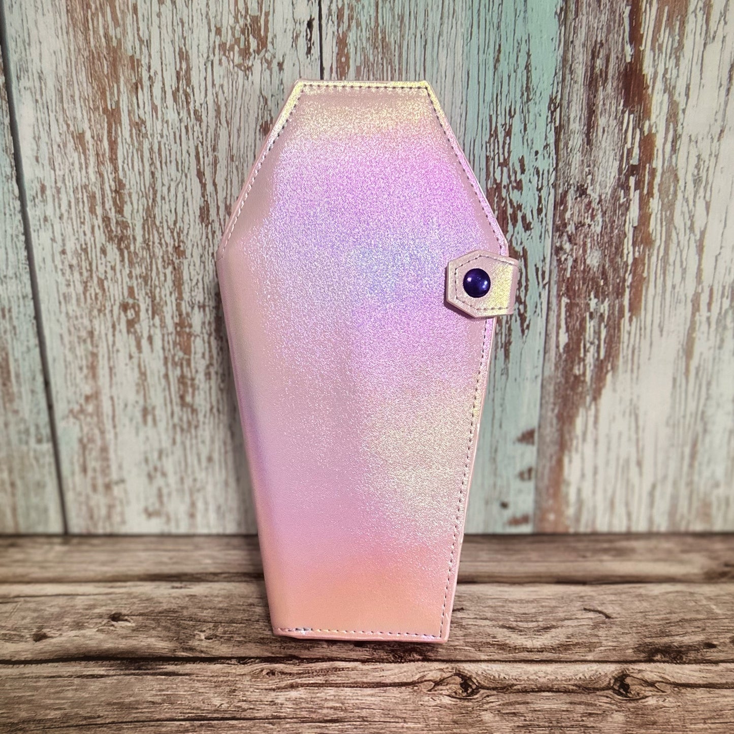 Front view of a tall handmade coffin-shaped wallet in shimmering iridescent vinyl that shifts between pink, lavender, and gold tones. The wallet features a matching coffin-shaped snap tab closure, fastened with a round rainbow-finished snap. Displayed upright against a rustic wood background.