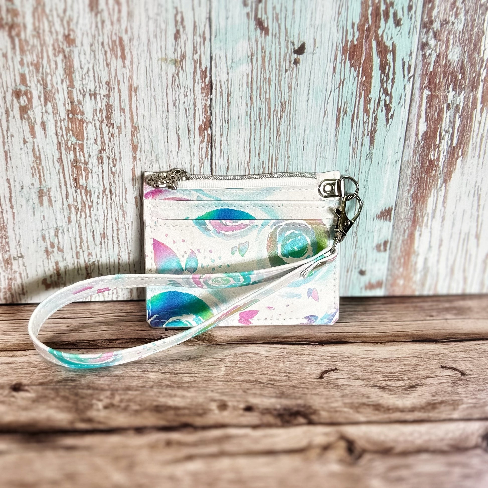 Handmade card holder with a detachable wristlet strap, featuring a pastel floral swirl print in teal, lavender, and pink on a white background. The wallet includes two card slots on the front and a top zipper closure with silver hardware. Displayed against a rustic wood backdrop.