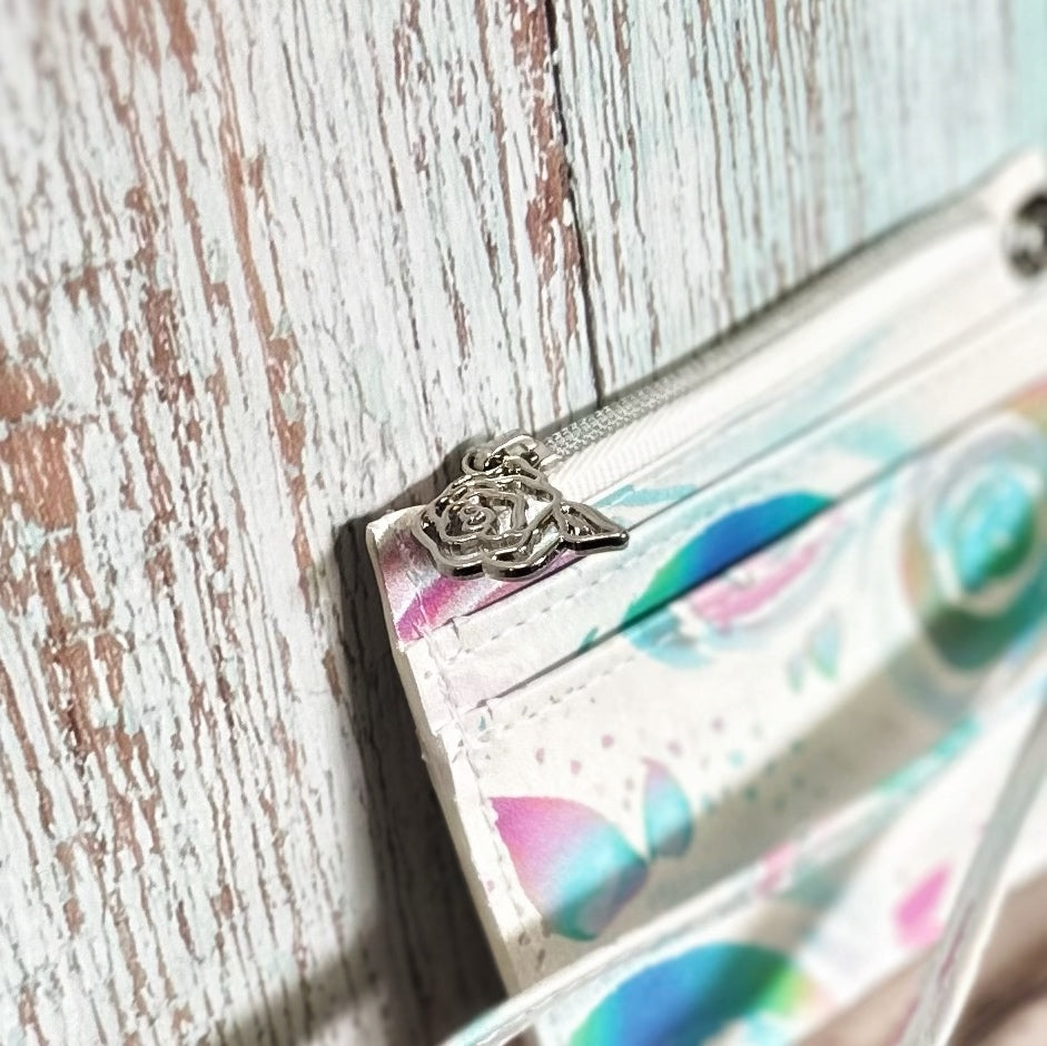 Close-up of the zipper pull on a handmade card holder, featuring a silver rose-shaped charm attached to a white zipper. The fabric shows part of the pastel floral swirl pattern in pink, teal, and lavender tones. Background includes distressed wood paneling.