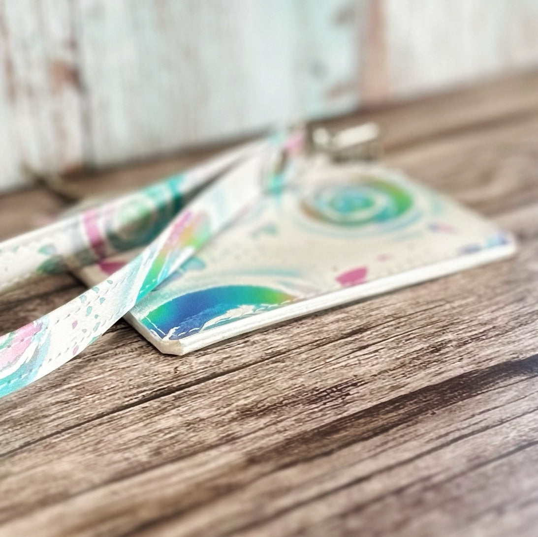 Angled close-up of a handmade card holder showing the neatly painted white edges and pastel floral swirl print. The attached wristlet strap matches the fabric pattern, and the card holder rests on a rustic wooden surface with a blurred background.