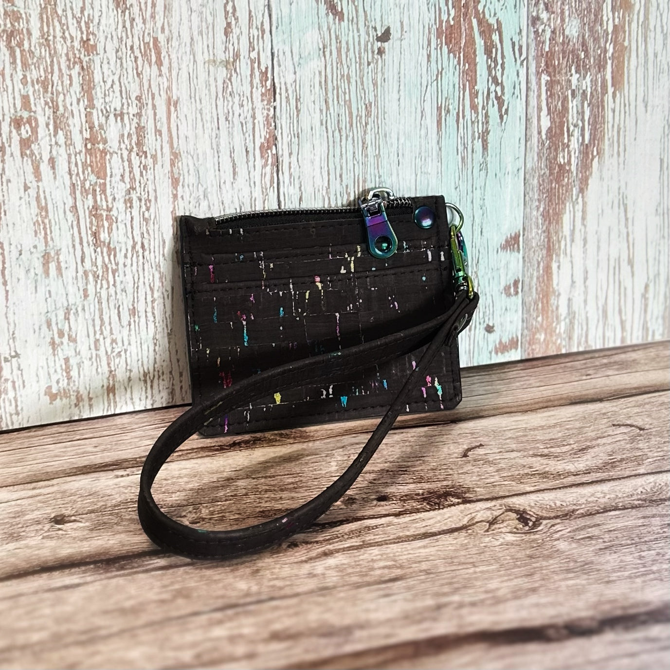 Handmade card holder with a detachable wristlet strap, crafted from black cork featuring subtle rainbow flecks throughout. The front includes two card slots and a top zipper closure. All hardware, including the zipper pull, d-ring, and clasp, is rainbow iridescent. Displayed upright on a rustic wooden surface.