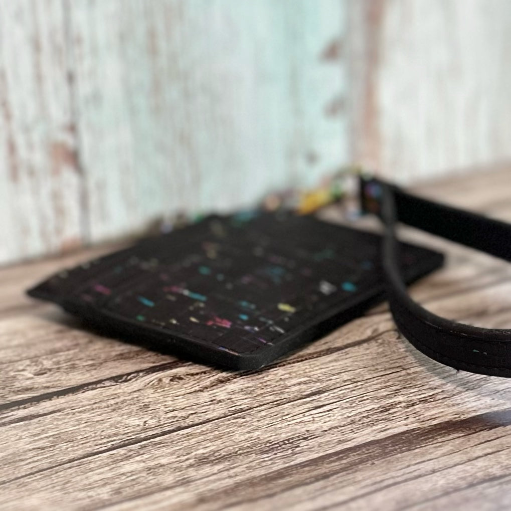 Angled close-up of a handmade card holder crafted from black cork with rainbow flecks, highlighting the matte black edge painting. The wristlet strap and rainbow hardware are partially visible in the background. Displayed flat on a rustic wooden surface with a blurred backdrop.