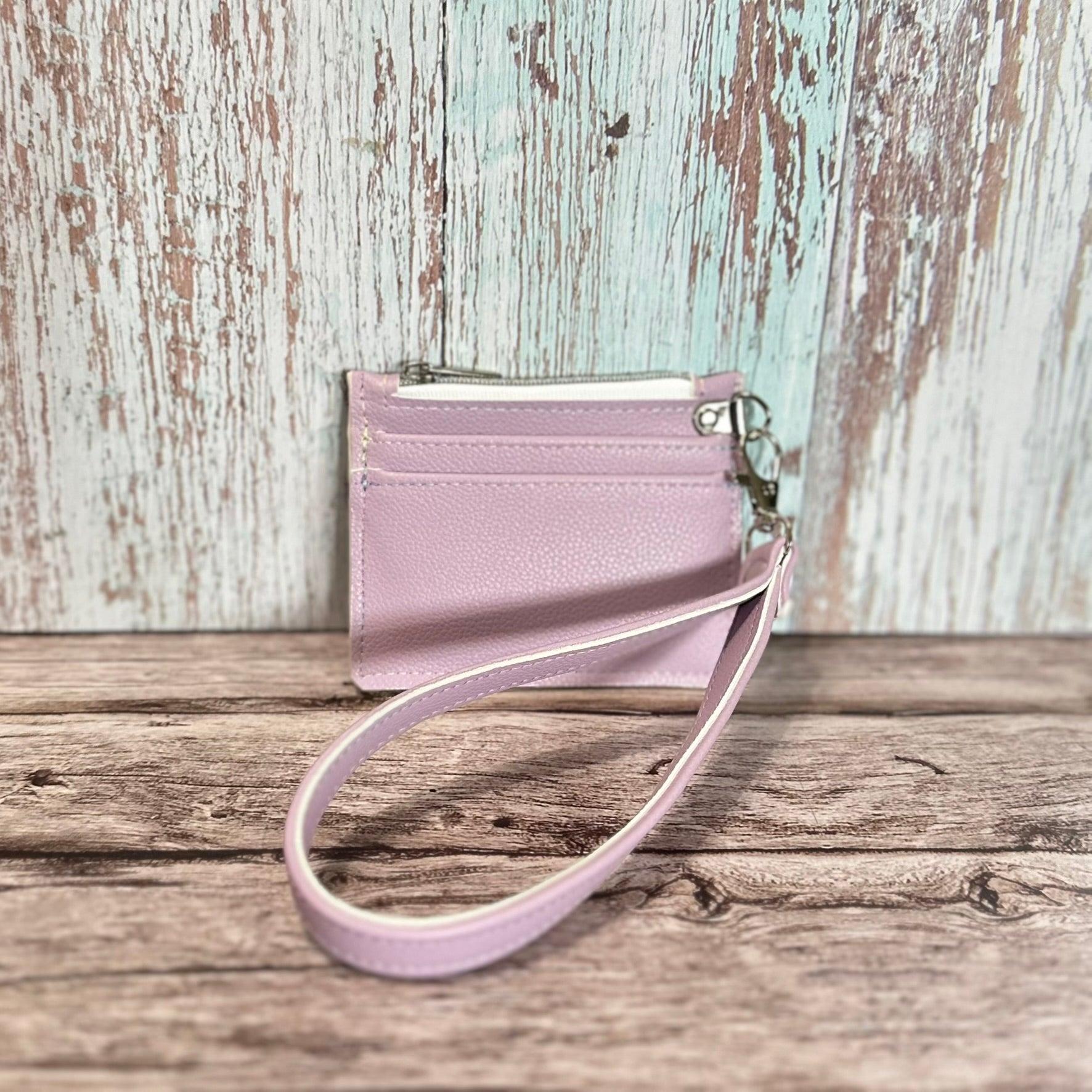 Handmade card holder with a detachable wristlet strap, crafted from lavender faux leather with a soft pebbled texture. The design includes two front card slots and a top zipper closure with silver hardware. Displayed upright against a rustic wood surface.