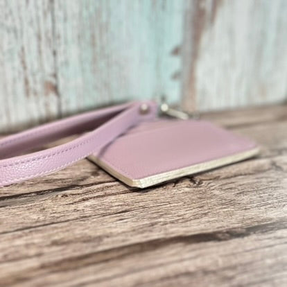 Angled close-up of a handmade card holder in lavender faux leather, highlighting the shimmer white edge painting along the base. The detachable wristlet strap is partially visible, and the card holder rests on a rustic wooden surface with a blurred background.