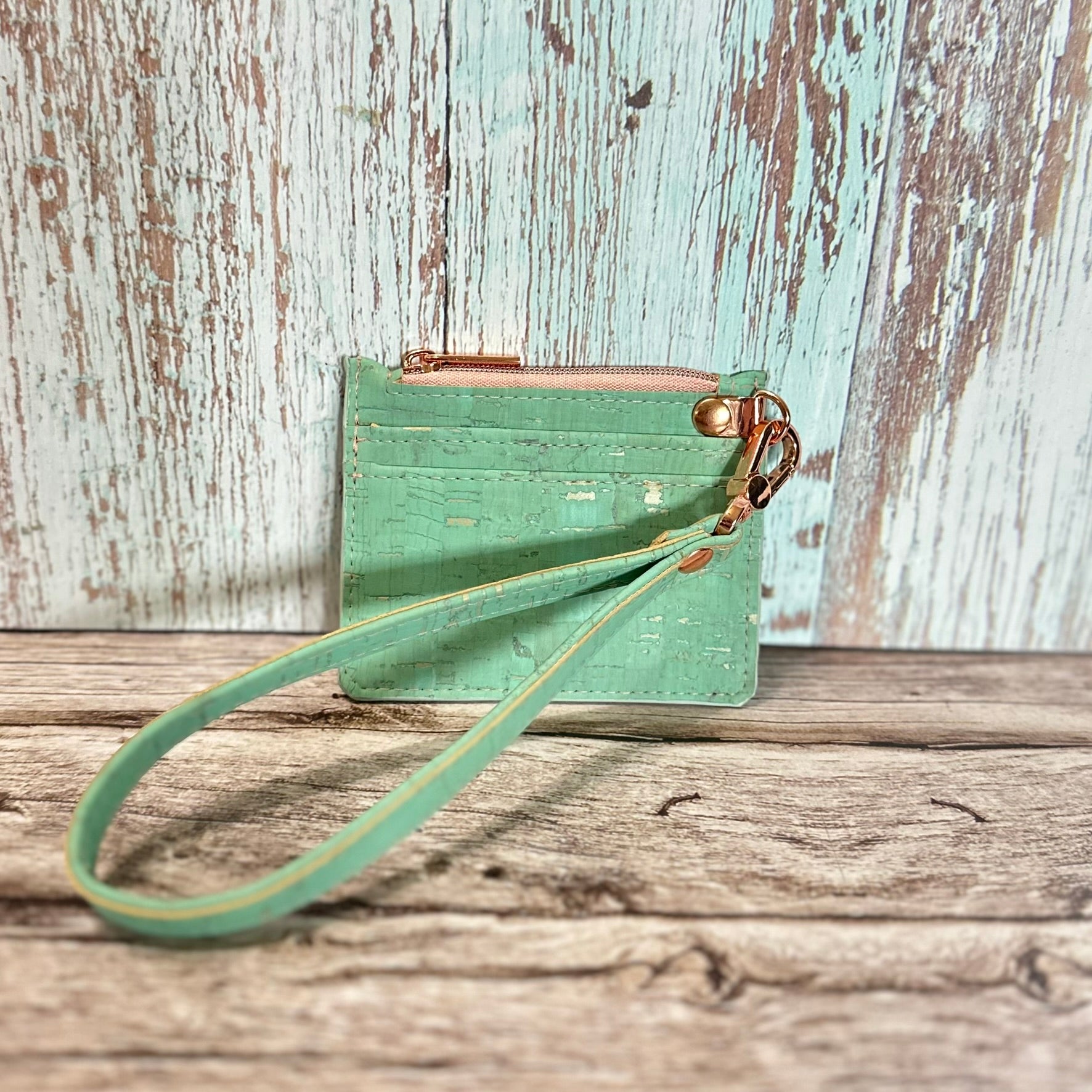 Handmade card holder with a detachable wristlet strap, crafted from mint green cork featuring subtle gold flecks. The front has two straight card slots and a top zipper closure in dusty rose, with rose gold hardware including the d-ring and swivel clasp. The wristlet strap matches the mint cork. Displayed upright on a rustic wood surface with a distressed turquoise and white background.