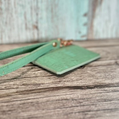 Angled close-up of a handmade card holder in mint green cork with subtle gold flecks, highlighting the smooth matte mint edge painting. The matching wristlet strap and rose gold hardware are partially visible, with the card holder resting flat on a rustic wooden surface against a softly blurred background.