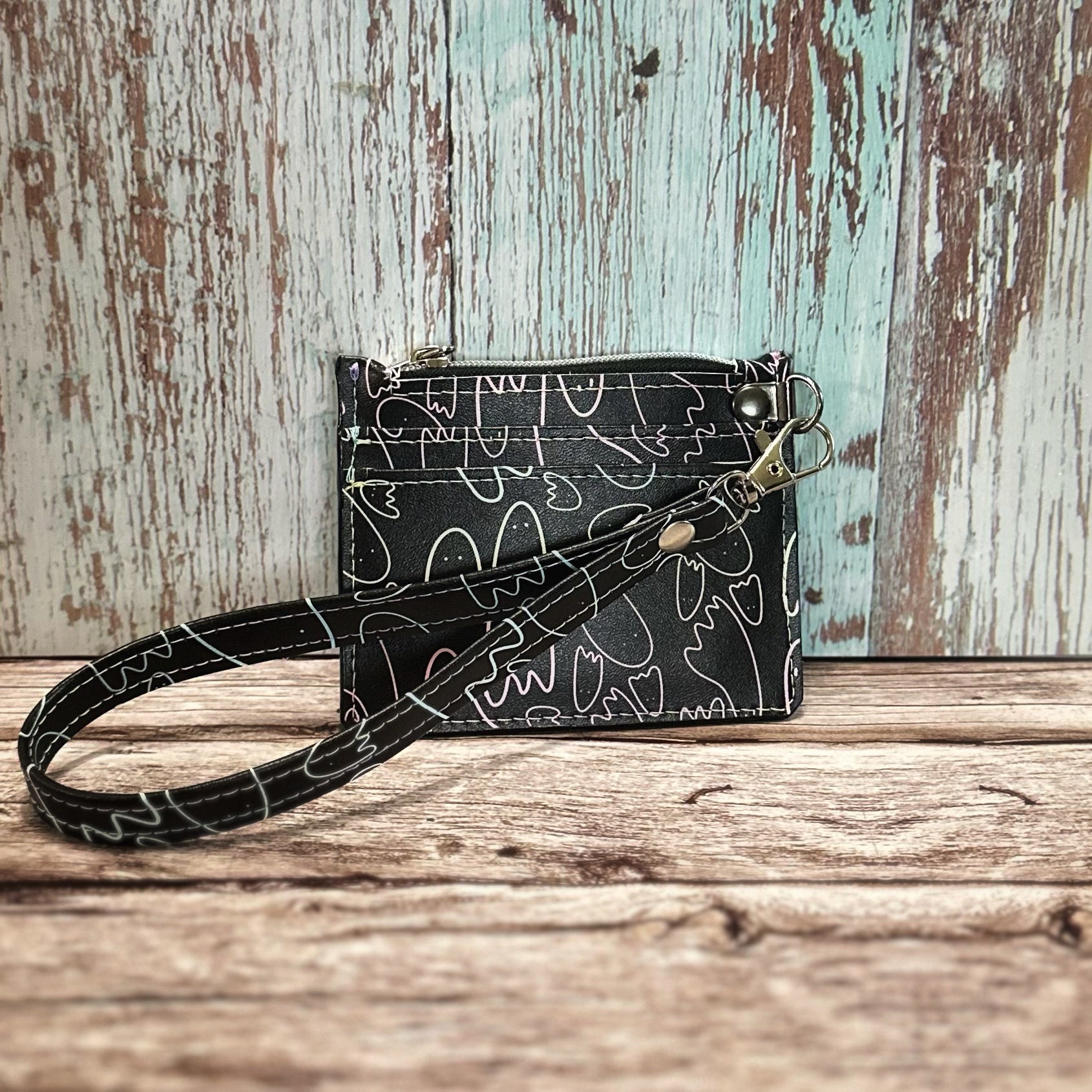Handmade card holder with a detachable wristlet strap, featuring a black background printed with whimsical ghost outlines in subtle pastel shades of pink, blue, green, and purple. Includes two straight front card slots, a top zipper closure, and silver hardware. Displayed upright on a rustic wooden surface with a distressed turquoise and white backdrop.