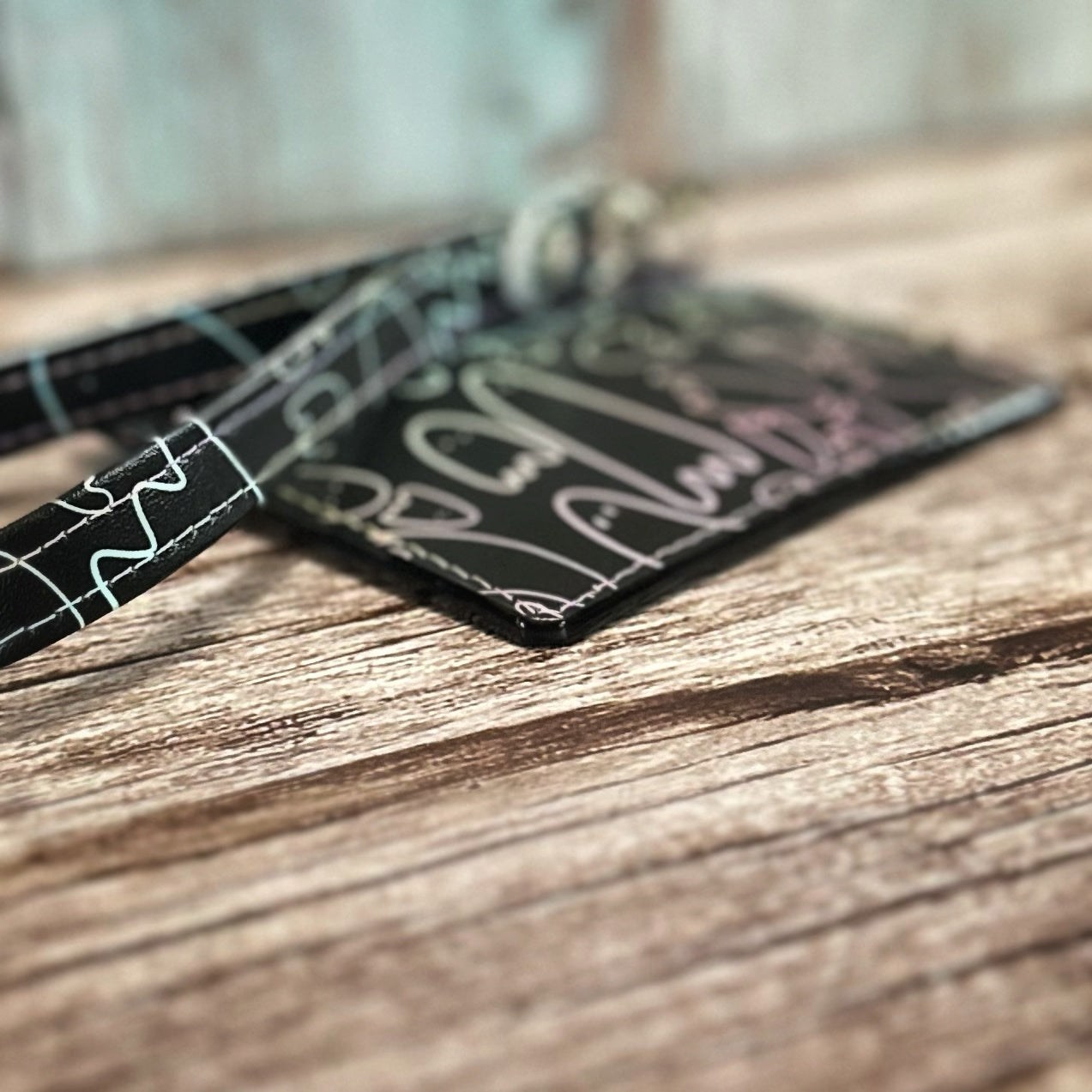Angled close-up of a handmade card holder with a black background and pastel ghost print, showing smooth black edge painting that matches the base material. The coordinating wristlet strap and silver hardware are partially visible. Displayed flat on a rustic wooden surface with a softly blurred background.