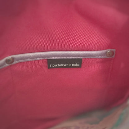 Close-up of the right slip pocket inside a handmade tote bag, made from bright pink fabric with lavender faux leather trim and metal rivets. A black fabric label is stitched into the pocket and reads, 'I took forever to make.'