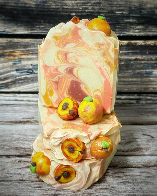 Stack of three handmade soap bars with a peach and cream swirl pattern. The tops are decorated with realistic soap embeds shaped like peaches and peach halves, featuring details like green leaves and brown pits. Set against a rustic wood background.
