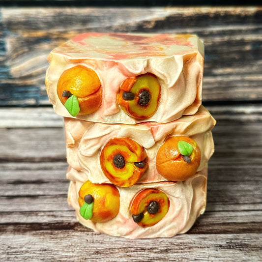 Close-up of three handmade soap bars stacked to show their tops. Each soap is topped with realistic peach-themed embeds, including whole and halved peaches with green leaves and dark brown pits. The tops have a textured swirl design in peach and cream tones.