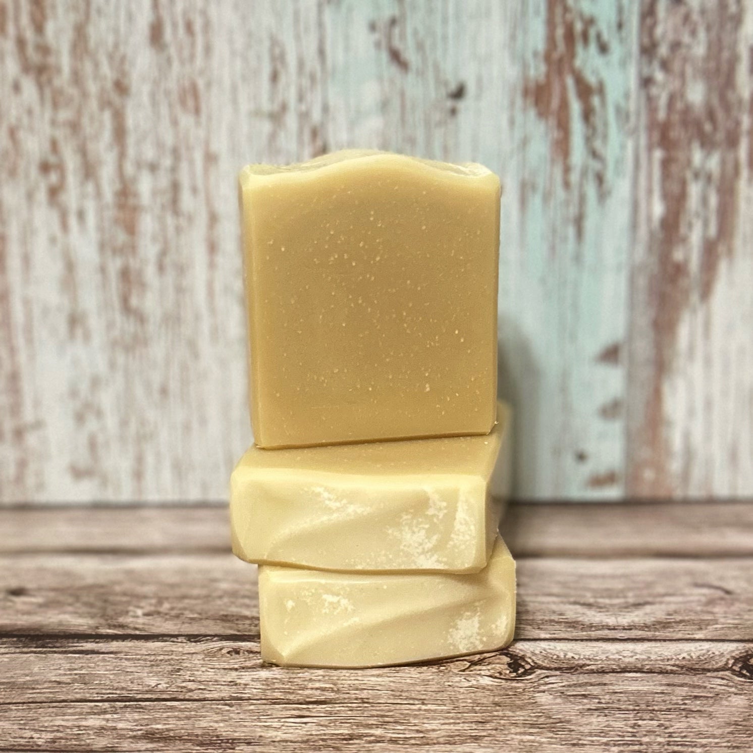 Three handmade soap bars in a warm, buttery yellow tone are stacked in a balanced arrangement. Two bars lie horizontally with gentle texture swirls, while the third stands upright with a softly curved top. The background is a rustic wood surface with a distressed pale teal and white wood panel backdrop.