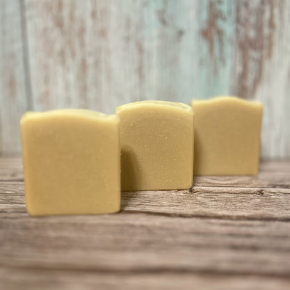 Three handmade soap bars in a soft, golden yellow tone are lined up diagonally on a rustic wooden surface. Each bar has a slightly curved top and a smooth, natural finish. The background features distressed pale teal and white wood paneling.