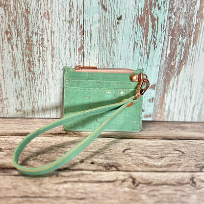 Handmade card holder with a detachable wristlet strap, crafted from mint green cork featuring subtle gold flecks. The front has two straight card slots and a top zipper closure in dusty rose, with rose gold hardware including the d-ring and swivel clasp. The wristlet strap matches the mint cork. Displayed upright on a rustic wood surface with a distressed turquoise and white background.